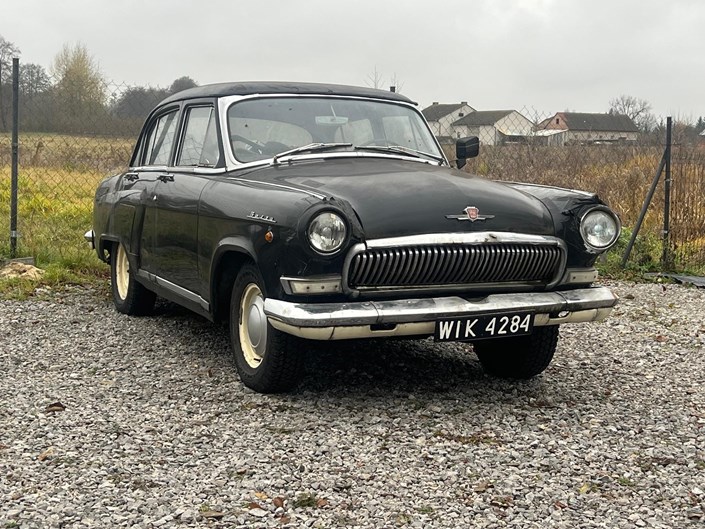 Zdjęcie przedstawia czarny klasyczny samochód GAZ-21 Wołga zaparkowany na żwirowym podłożu w otoczeniu wiejskiego krajobrazu. Auto ma chromowany grill, okrągłe reflektory i charakterystyczne emblematy na masce oraz błotnikach.